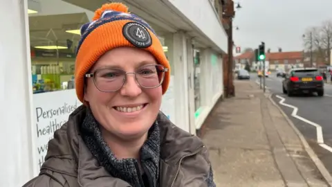 Andrew Turner/BBC Jessica Murison is wearing a brown jacket and grey mottled jersey, and an orange and blue hat, featuring a black logo badge. She is standing outside the Acle Pharmacy on the high street. She is wearing glasses and is smiling.