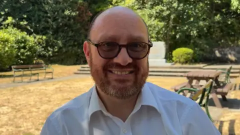 A man with very short hair on the side of his head and a brown and grey beard. He is wearing sunglasses and smiling at the camera. He is wearing a white shirt. Behind him, park benches and trees are visible.