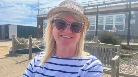 Richard Daniel/BBC Pauline Turner has blonde shoulder-length hair and is wearing a hat and sunglasses. She is wearing a navy and white striped top and is smiling.