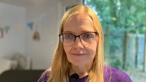 Shariqua Ahmed/BBC Michelle, a woman with blonde hair and glasses. She is wearing a purple T-shirt and is looking into the camera.