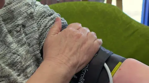 A close-up of a black blood pressure monitor strapped around an upper arm. A hand is resting on the monitor. The person cannot be seen in full but is wearing a grey jumper.
