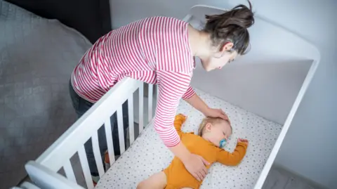 A woman leaning over a white cot, adjusting a baby lying on their back on a light-patterned mattress. The adult is wearing a red-and-white striped long-sleeved top, and the baby is dressed in an orange outfit. The scene is indoors, with a bed visible beside the cot.