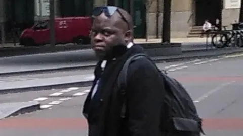 Simon Levy wearing dark clothing walks along a city street with a backpack, with buildings, parked vehicles and bicycles visible in the background.