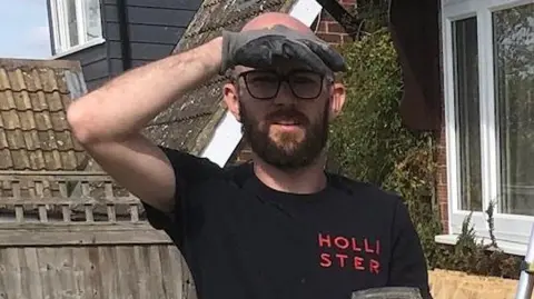 Dervin family Greg is using a hand to cover his eyes from the sun while garderning. He is wearing a black T-shirt.