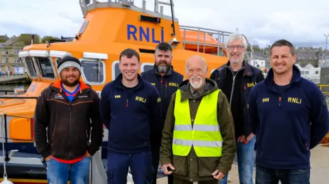 Lewis Lifeboat Five men wearing RNLI fleeces and Derek Adams with short white hair and beard wearing a dark green waterproof coat and yellow hi-vis jacket, standing in a row. Behind them is an orange RNLI lifeboat moored in a harbour.