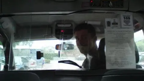 Barry Cook Barry Cook, pictured here in the 2000s in his taxi at the time. He is sat in the driver's seat and is looking over his shoulder at the camera through the plastic partition. 