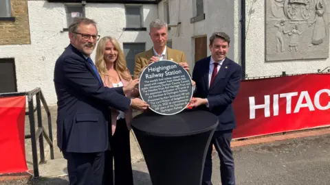 A man stands next to a woman and in the centre is Niall Hammond and next to him is Alan Strickland. They are all holding a blue circular plaque that reads 'Heighington Railway Station'.