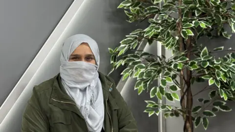 School of Coding and AI Shakielah Bibi is sat on a chair by a tall, green plant and some stairs. She has her face and head partially covered by a scarf with a green overcoat on.
