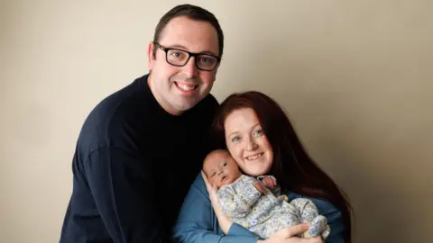 A man in a black jumper- hugging mum Georgina, who has reddish hair and is wearing a blue top and holding baby Robyn. She is wearing a floral baby-grow.