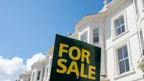 Getty Images A black sign with 'for sale' in yellow writing printed on it, in front of a white house.