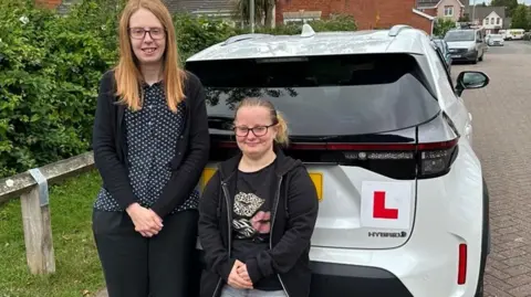 BBC Chelsea Moon, who has a bone condition that stunts growth, waited a year for lessons with instructor Emma Hallett (left). Chelsea is just over four feet tall and is wearing a black hoodie, grey jeans and glasses. Emma, also in glasses, is wearing a black cardigan trousers and polka dotted shirt. They are standing behind a white car with learner plates.