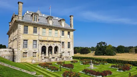 An old building, which looks part of a stately home, stands proudly in front of gardens on a clear and sunny day.