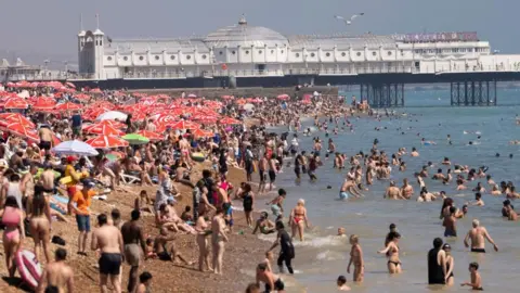 A busy beach which is full. People sit under red umbrellas, as they watch peoples swimming in the murky sea. There's a large pier in the background