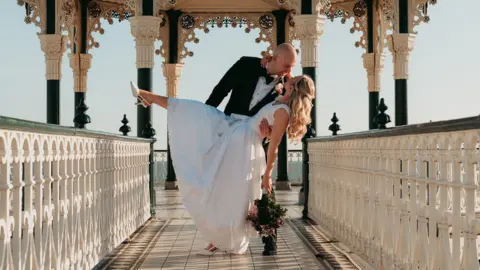 The image shows a groom holding his bride as she dips with one foot in the air and a bouquet in her hand. They are on what looks like a pier and the sky behind them is clear.