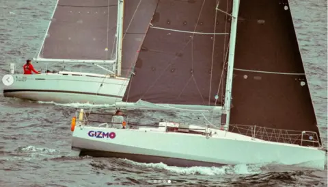 Meg Niblett A yacht with the name Gizmo on the starboard side and a woman at the helm with dark sails and another yacht behind with a skipper in a red top.