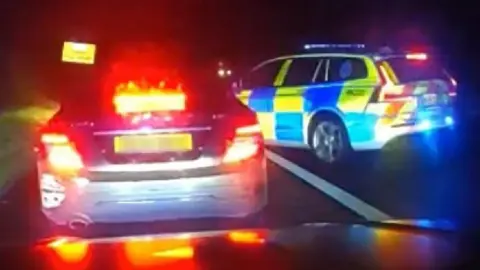 A police car pulls in front of a black car in the dark on the road. It looks like the car has been boxed in to be stopped for checks.