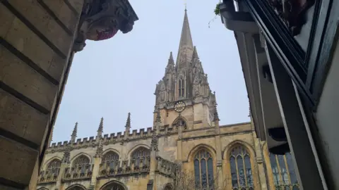 A gothic church in typical Oxford yellow/grey colours, with a tall spire adorned with a clock.