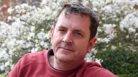 Stephen Davison A man with dark hair looks at the camera. He is wearing a red jumper, and behind him there is a bush with white flowers.