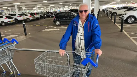 John Devine/BBC Tony Johnson is smiling at the camera while standing in a large car park. He has a supermarket trolley with him. He has short grey hair and is wearing a blue jacket over a white and black patterned shirt. There are lots of cars parked in the background.