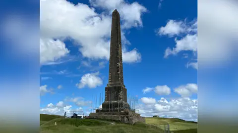 A tall stone monument on a grassy hillside set against a blue sky with some small white clouds