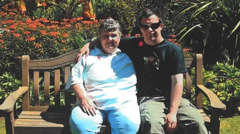 Handout A woman with a white top and pale trousers is sitting on a brown bench. The man next to her has a black T-shirt with an image on it, khaki trousers and is wearing sunglasses. He has his arm around the woman. The bench is in front of a flower bed.
