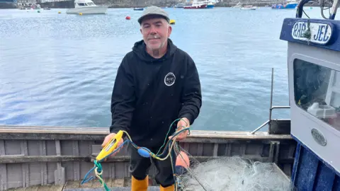 Will Shugg stands on his boat holding a pinger, which is yellow and shaped like. a banana, and wearing a flat cap and a black hooded jumper