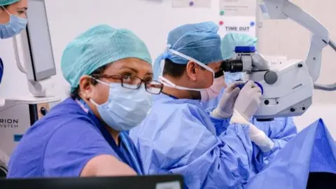 A cataract operation taking place at Sunderland Eye Infirmary. Four medical staff with face masks, scrubs and gloves have surrounded an operating table. One of the clinicians is looking through a machine. 
