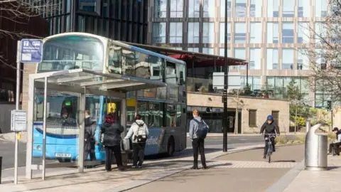 Four people queue to get on a bus on Charter Row in Sheffield city centre, with a cyclist riding down the cycle path towards the camera. A silver bin is to the right with a person sitting on a bench in a black cap next to a tree
