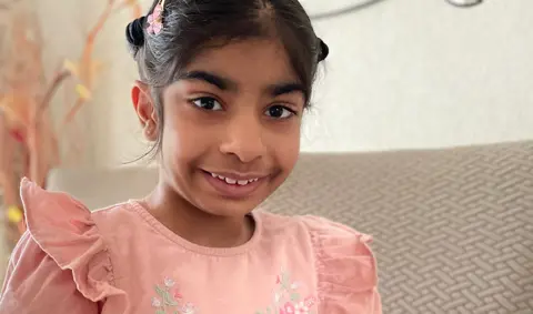 Eight-year-old Kriti in a pink dress sitting on a sofa in the family's house