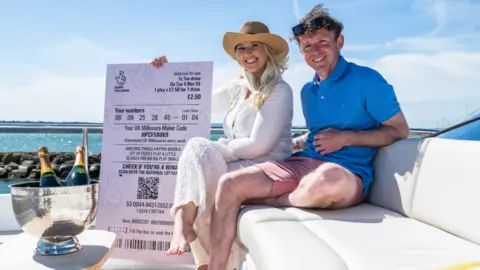 A man and a woman sat side-by-side on a boat holding a huge EuroMillions receipt 