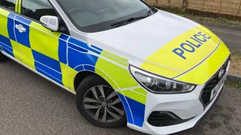 BBC A Devon and Cornwall Police car parked on a residential street. It has blue and yellow squares across its body. The rest of the car is white and has the word "police" written in blue block capitals on a yellow background on its bonnet.