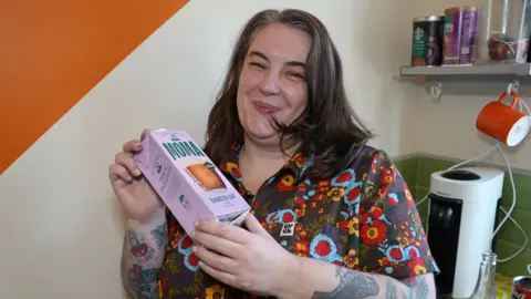 Louise Brown smiling and looking down the camera. She is standing inside a kitchen area; there is equipment on the counter behind her and an orange mug hanging from a hook under a shelf on the wall on the right of the image. There are cans of coffee on top of the shelf. Louise has straight, shoulder-length hair, that is curling around her cheeks and chin. She is wearing a heavily patterned, flowery top and is holding a carton of 'barista oat' drink.