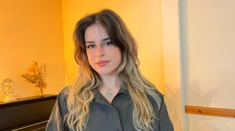 BBC a woman smiling at the camera in front of a mantelpiece. She is wearing a grey shirt