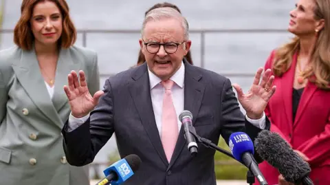 AFP via Getty Images Australia's Prime Minister Anthony Albanese (C), wearing a dark suit with a white shirt and pink tie, gestures with his hands as he speaks into three news channel mikes. On his left in the background is Minister for Communications Anika Wells