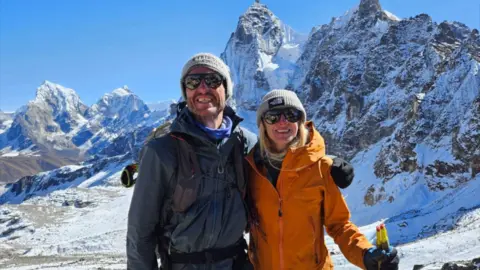 Rik de Louche Rik and Liv have their arms around each other as they smile for a photo in front of a mountain with snow in the background. Liv is wearing a beanie hat, sunglasses and a orange coat and is holding hiking polies. Rik also has a beanie hat on and a dark blue coat.