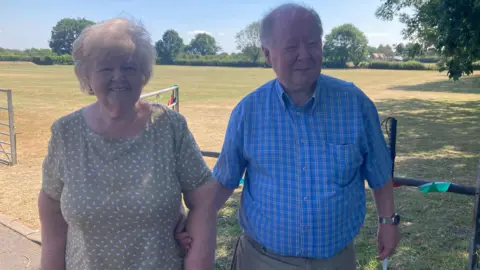Chris Duggan and his mum Beryl, standing in front of a field on a sunny day. Beryl is wearing a sage green dress with a white pattern on it and standing on the left of the image. Chris has his arm looped through hers and is wearing a blue checkered shirt. He is holding a white cane.