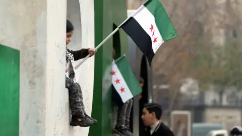 A child sits on the windowsill waving the new Syrian flag.