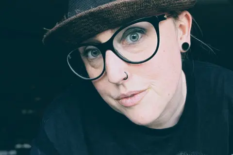 Kate Quinn Kate Quinn wears a dark had a thick-rimmed glasses. She has blue eyes and a nose piercing and a large earring in one ear. 