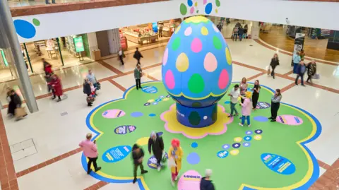 Merry Hill Shopping Centre The blue, green, pink and yellow egg stands in a large space in the shopping centre with people looking at it or walking by. The floor is stands on is also painted green, blue yellow and pink.