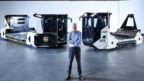 A man standing an a dark warehouse that has dim lighting. He is standing with arms crossed in front of two self-driving vehicles that are used to move airport baggage. He is wearing suit trousers and a blue shirt and has short grey hair. He is smiling. 