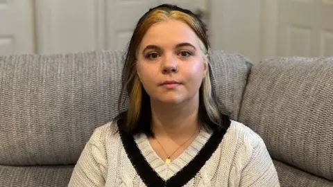 A woman sitting on a sofa with dark, shoulder-length hair that has a blonde streak. She also has a nose ring and is wearing a white V-neck jumper with a black V.