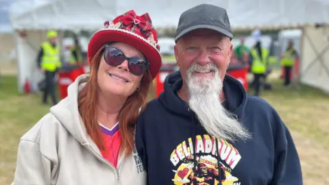 Retired distiller Trish McCarroll, 61, and retired lorry driver Eddie McCarroll, 72, from Methil, are standing outdoors at the festival site. Trish has shoulder length red hair and is wearing a red bowler hat. Eddie has a long white beard and is wearing a grey cap and a Belladrum branded hooded top.