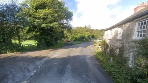 Google A google street view of a crossroads. On the right there is a stone house and on the left a gated green field, with tall trees. 