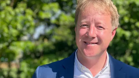 Paul Bristow is standing outside on a sunny day in front of a green bush. He is smiling at the camera while wearing a blue suit jacket and white shirt. He has blond hair.