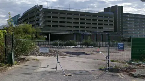 Google The Greyfriars site in Northampton, with metal gates across the entrance. A concrete surface is visible behind the gates, and a multi-storey car parking in brick and concrete is beyond. The sky is cloudy and grey.