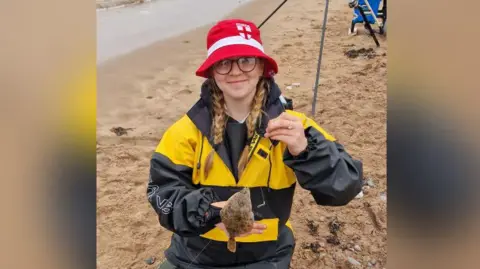 Family handout Lexie wearing yellow and black coat while posing with a small turbot fish