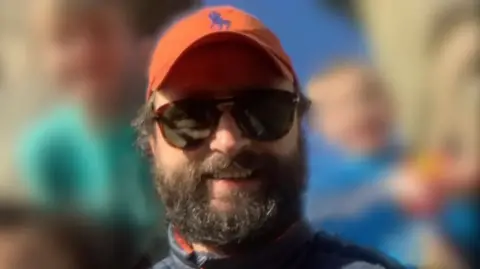 Family photograph Carl O'Keeffe is smiling brightly at the camera. He has brown hair and a bushy brown beard. He is wearing sunglasses and an orange cap. The people behind him are blurred out.