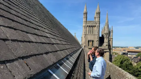 Peterborough Cathedral Two men in blue shirts are looking at tiling on top of part of a roof of a cathedral. Towers can be seen in the background against a clear blue sky.