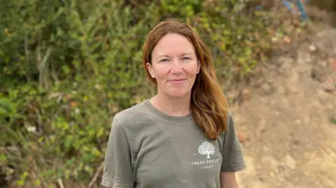 BBC Alex Morel smiling at the camera. She has brown hair and is wearing a green T-shirt. She's stood in front of bushes.