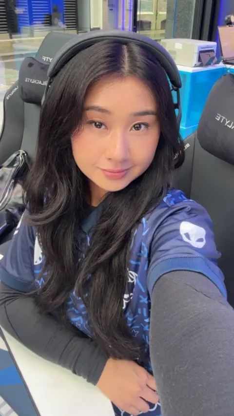 @karagii A woman wearing a gaming jersey and sitting in a black gaming chair. The person has long dark hair and is wearing a headset. In the background, there is a blue-lit gaming setup with a monitor, keyboard, and other equipment on a desk near a glass wall.